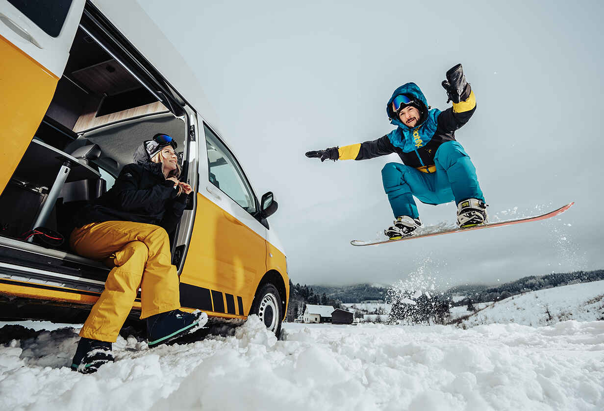 muž skákající na snowboardu s ženou ve žlutém autě, která se na něj dívá
